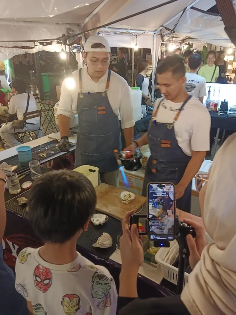 Two Ocean Tide Ventures shuckers working the counter under a torch flame while a customer films the stall on a phone