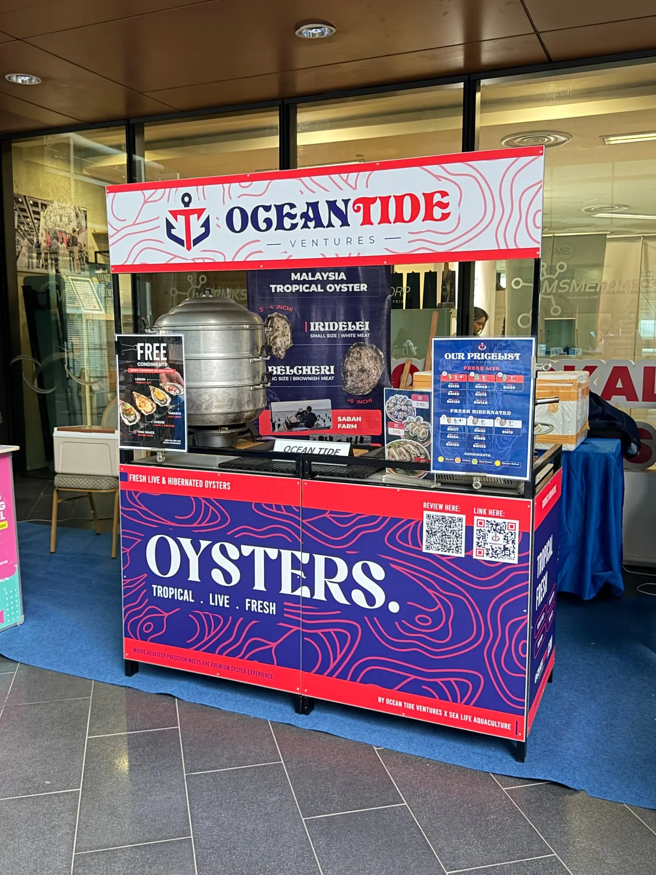The Ocean Tide Ventures oyster cart with its banner, pricelist board and tall aluminium steamer, standing on blue carpet indoors