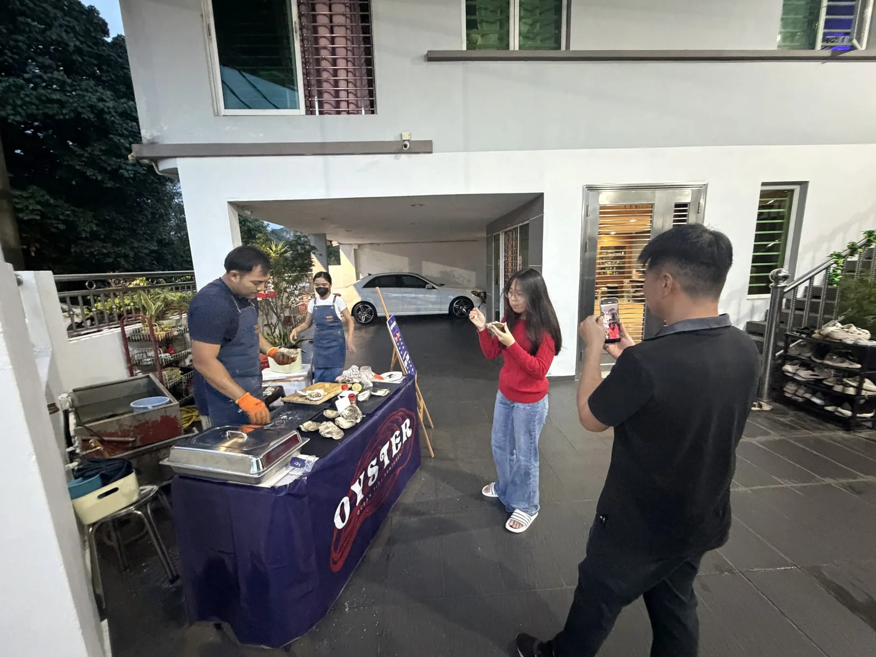 An evening pop-up oyster table with a purple Ocean Tide Ventures skirt, one person shucking while a guest tastes an oyster from the shell