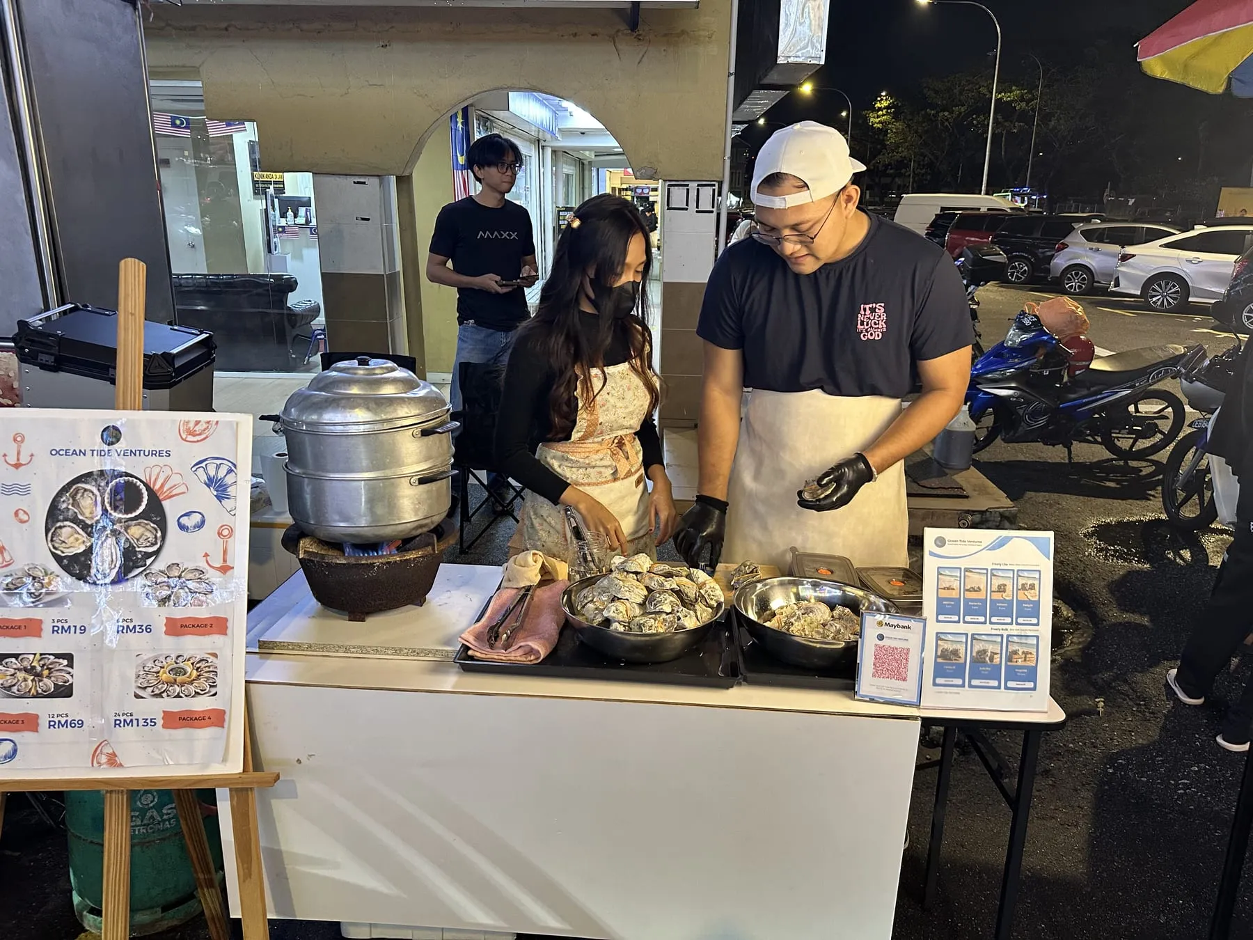 Two people in aprons and cut resistant gloves shucking oysters at a night stall counter, a tall aluminium steamer on a gas burner beside them