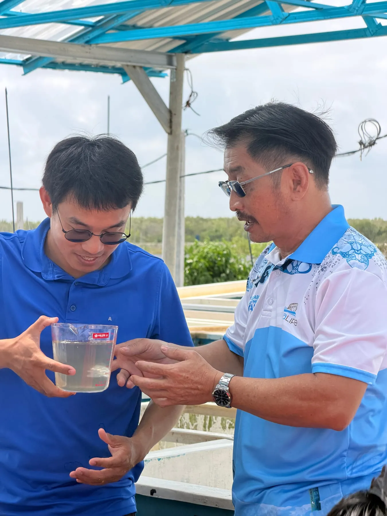 Two men under a blue roofed shelter examining a clear jug of cloudy pond water at a coastal aquaculture site